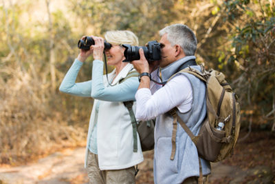 Image d'un couple âgé regardant vers la gauche ; la fille est derrière le garçon et a des jumelles, le garçon utilise un appareil photo.
