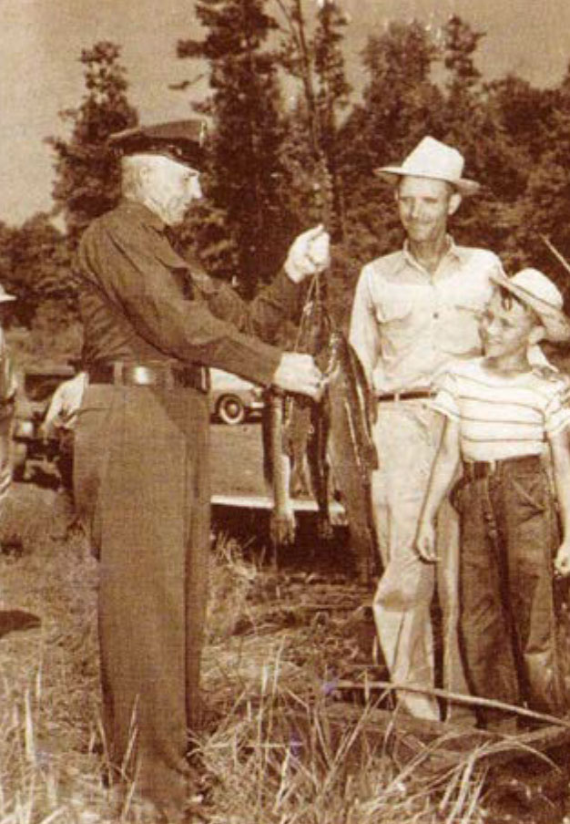 Image en noir et blanc d'un garde-chasse tenant un poisson et parlant à deux personnes.
