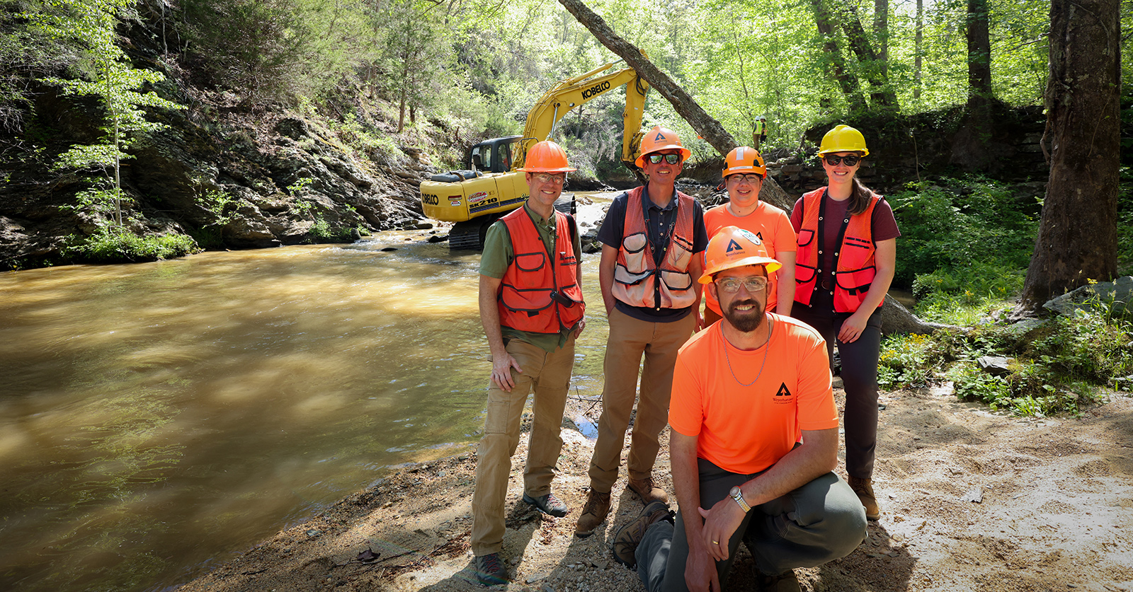 Une photo de cinq personnes en gilet orange et casque de protection posant devant une pelleteuse dans une rivière peu profonde.