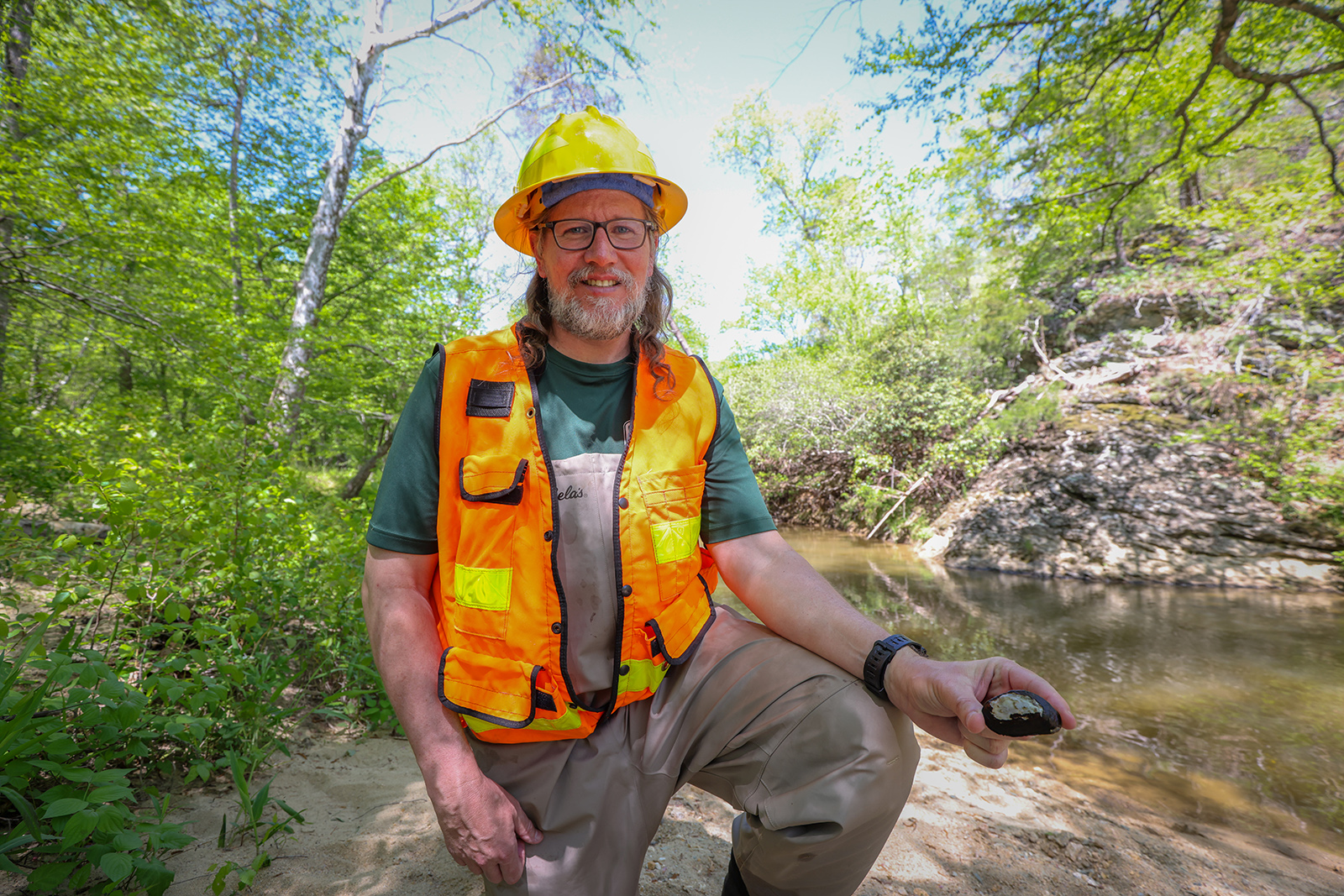 Un homme portant un casque de protection et un gilet orange pose avec une moule à épinards près d'un ruisseau en été avec des arbres verdoyants.
