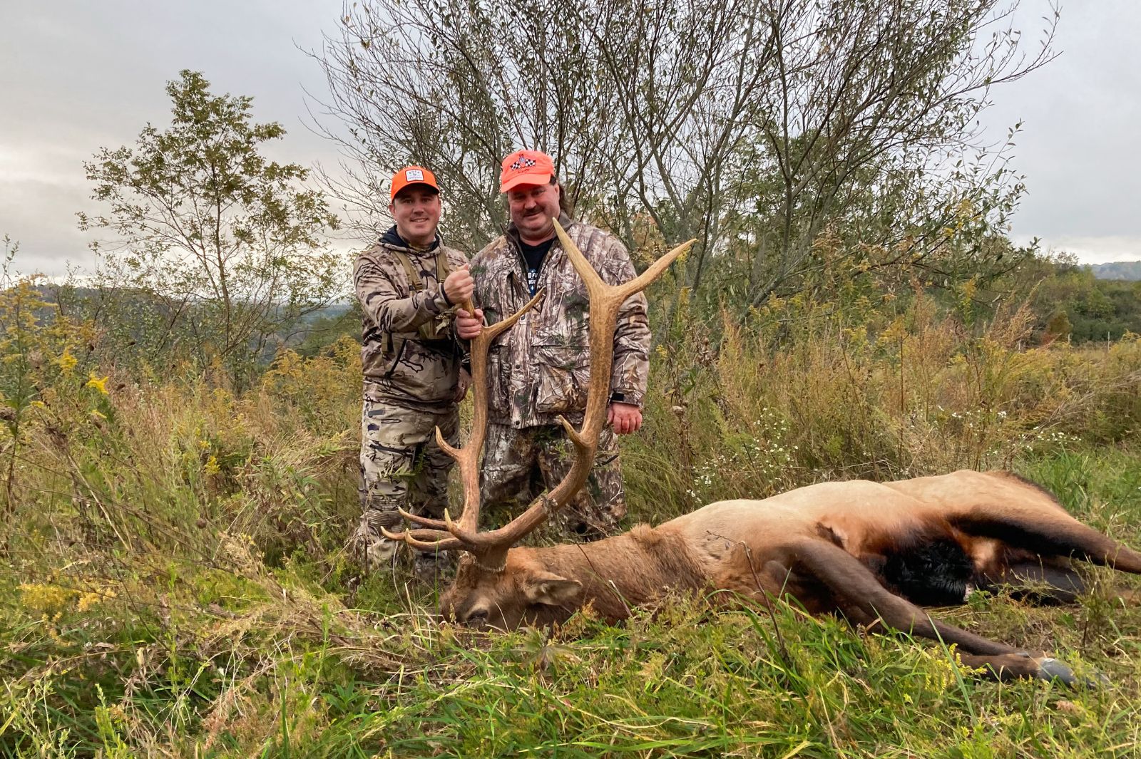 Une photo de deux chasseurs en camouflage et blaze orange posant avec un grand élan mâle couché sur le sol.