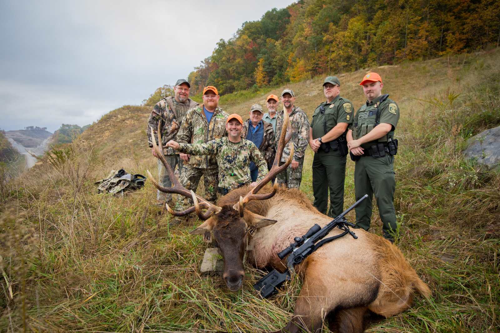 Une photo prise de six chasseurs en camouflage et orange vif posant avec un grand élan mâle couché sur le sol et deux agents de la police de la conservation en uniforme.