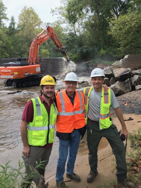 Construction d'un barrage avec un tracteur-pelleteuse démolissant le barrage au fond à gauche avec trois hommes (Alan Weaver, coordinateur du passage des poissons en Virginie, et les partenaires de l'agence) qui se tiennent ensemble en souriant pour une photo. 