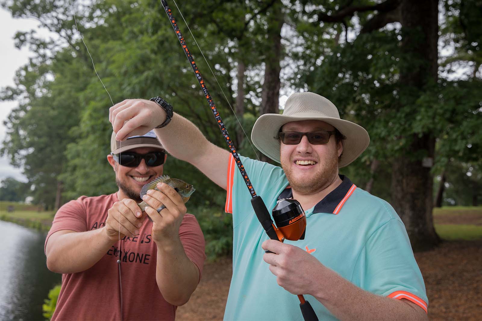 Deux hommes au bord d'un lac. Une personne tient une canne à pêche tandis que l'autre tient un poisson qui a été retiré de l'hameçon.