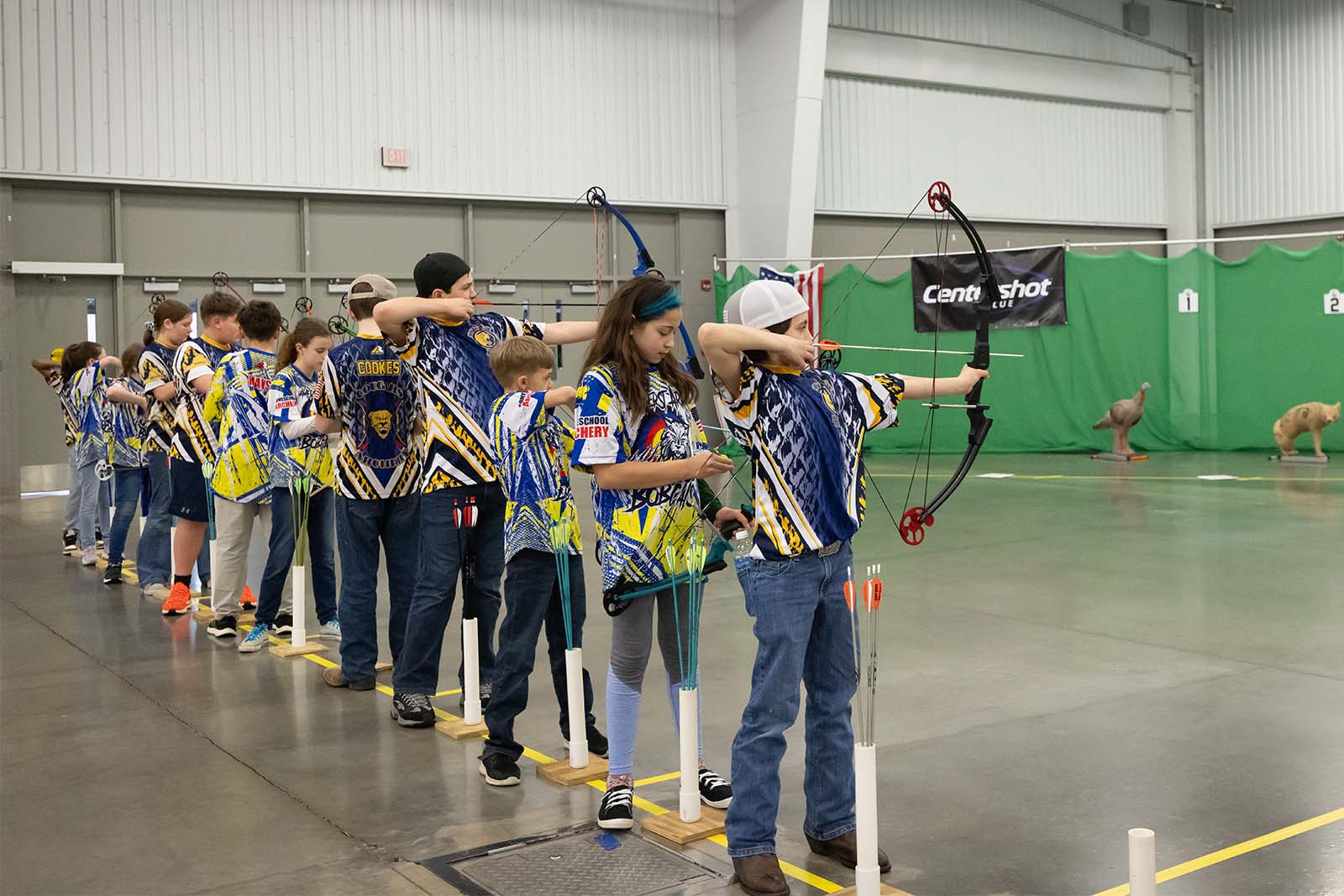 Les jeunes archers se tiennent en ligne avec leurs arcs, prêts à tirer.
