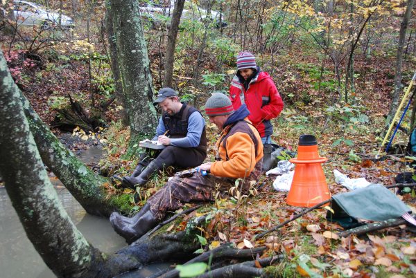Un trio de biologistes assis près d'un cours d'eau enregistrant les données sur les tortues des bois qu'ils ont recueillies au cours de leur étude.