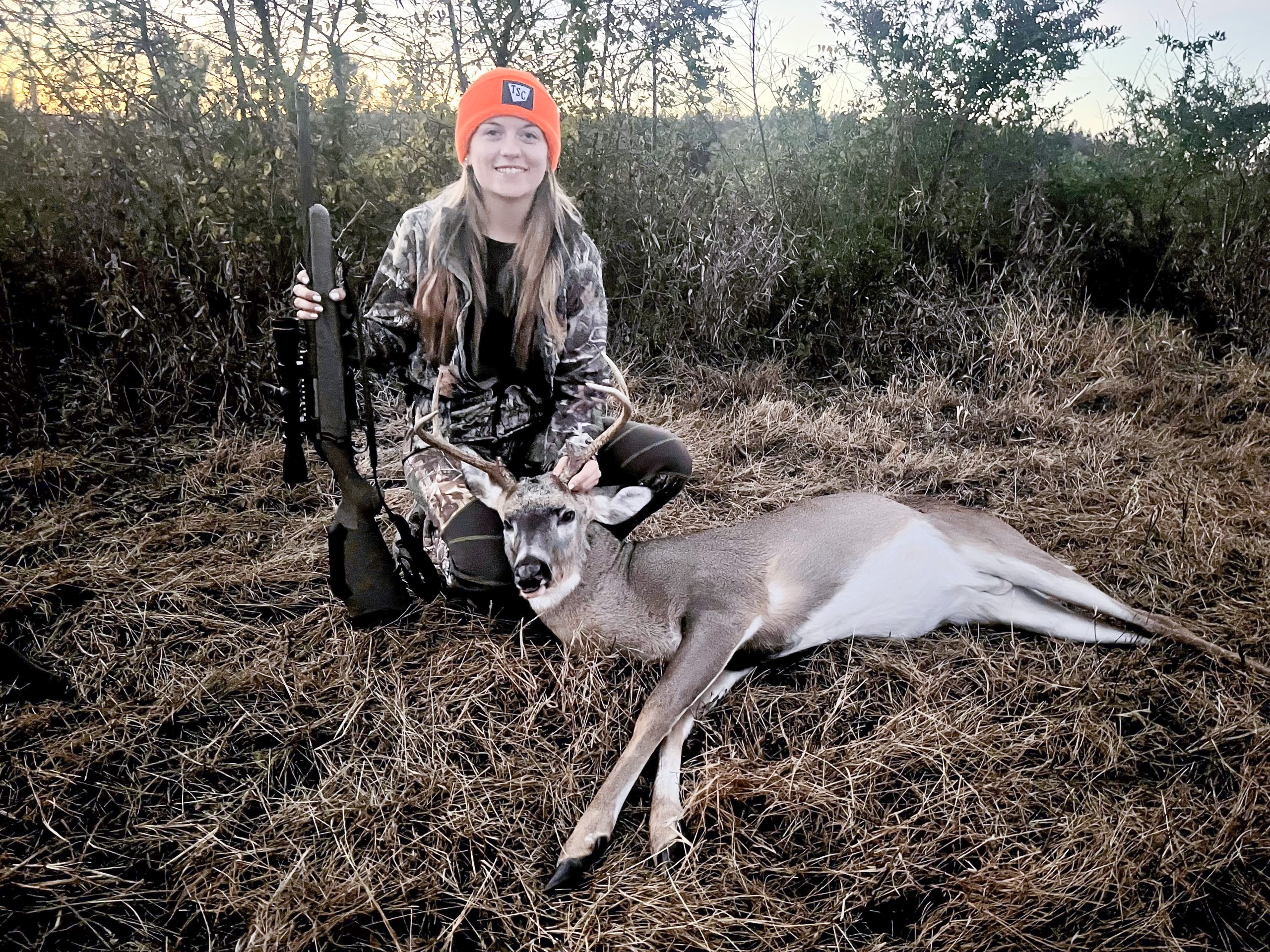 Photo d'une jeune femme tenant un fusil et agenouillée derrière un cerf abattu.