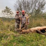 Deux hommes posant avec un élan mort dans un champ d'herbes hautes.