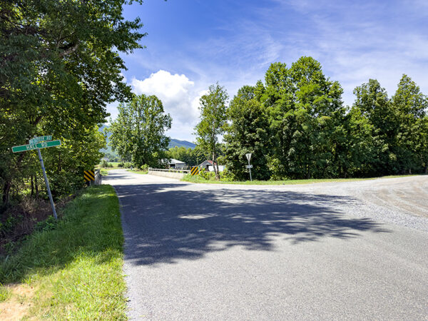 Vue pittoresque d'une intersection rurale avec une route de gravier qui bifurque vers la droite. Des arbres verdoyants bordent les deux côtés de la route sous un ciel bleu clair, avec quelques nuages visibles. Une plaque de rue indique la direction d'une route proche, et une vue lointaine de montagnes est visible à l'arrière-plan.