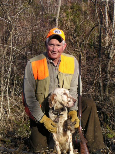 Image d'un homme portant un gilet de visibilité orange et un chapeau à côté de son setter anglais Gracie tenant une bécasse des bois du jour de Noël.