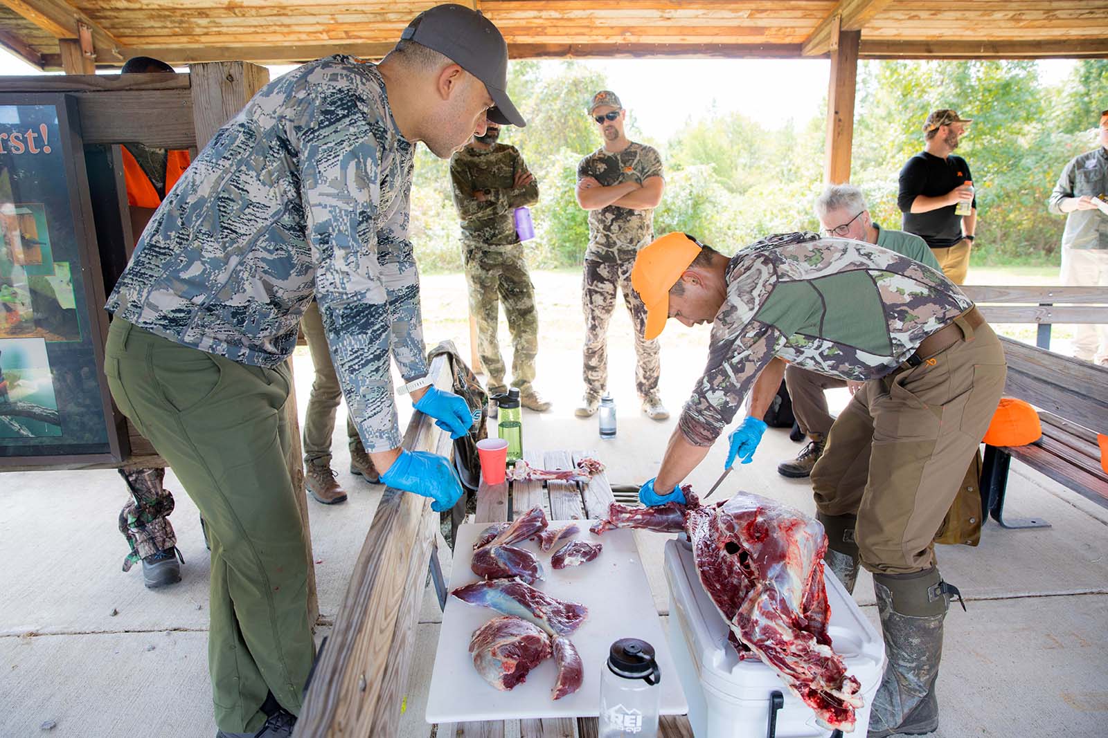 Photo d'un homme dépeçant un cerf sur une table, sous le regard attentif d'autres personnes. 