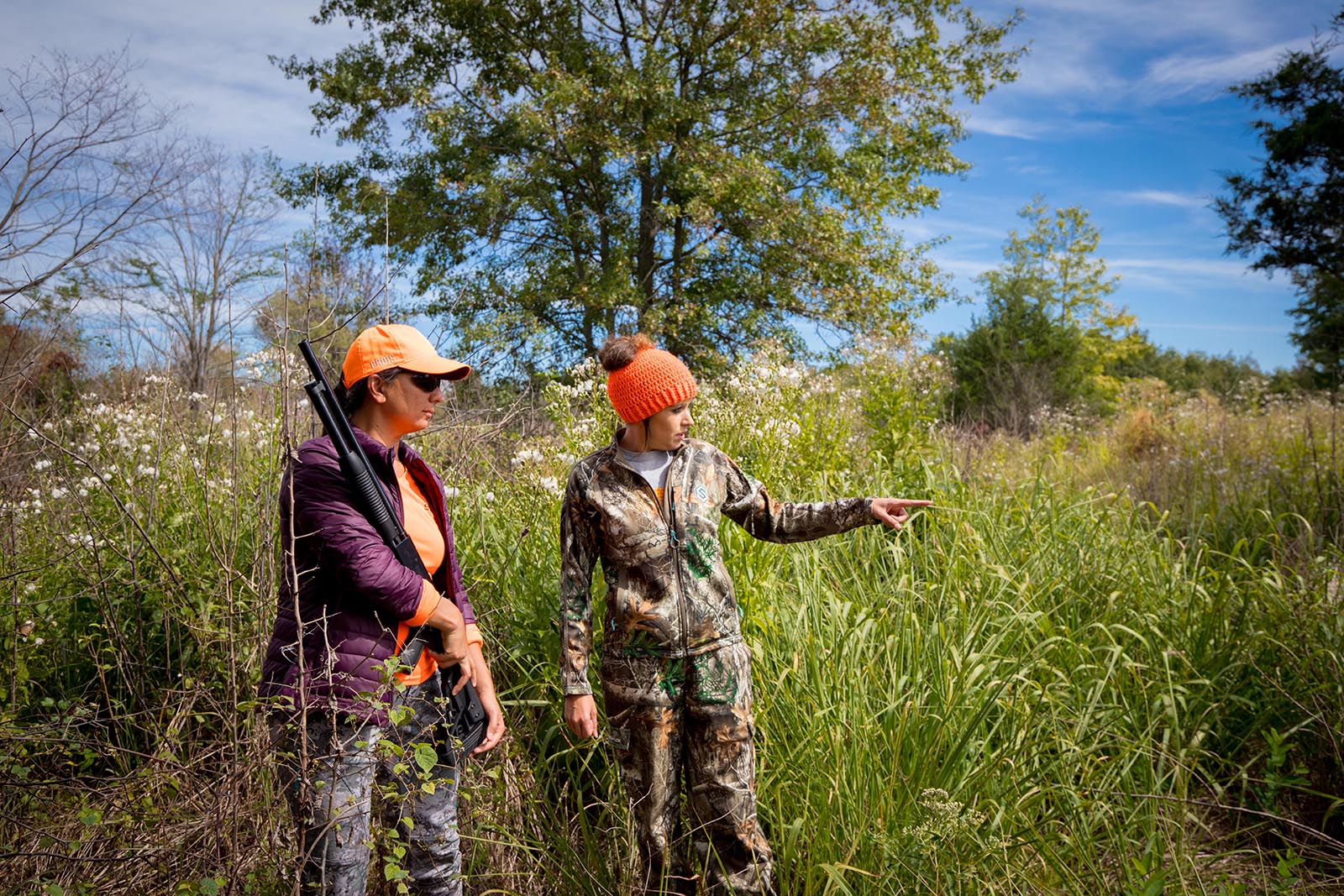 Une photo de deux femmes en tenue de camouflage et en orange vif, debout dans un champ, l'une dirigeant la femme qui tient un fusil.