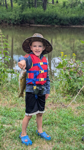 Un jeune garçon, âgé de 5, avec un chapeau dans un gilet, pose en tenant le poisson par la bouche.