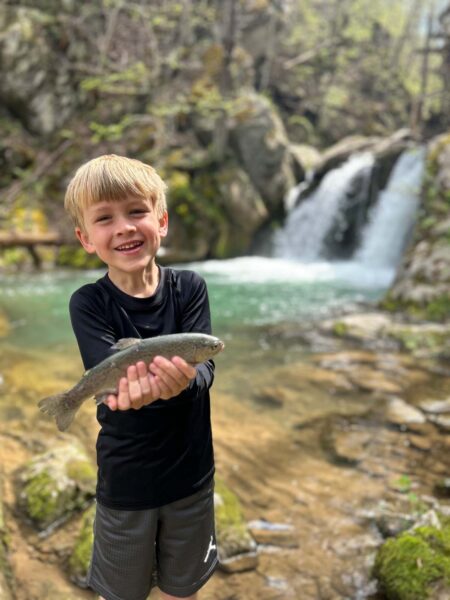 Un petit garçon souriant tenant un poisson pour l'appareil photo devant une cascade.