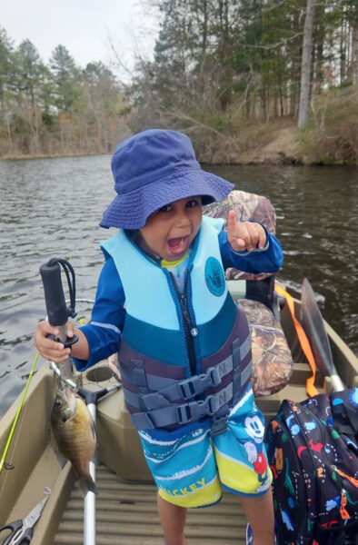 Un petit garçon très excité, portant un chapeau à godets, sur un bateau, tenant un poisson.