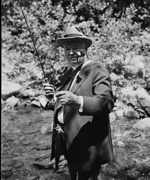 Photo en noir et blanc du président Herbert Hoover, vêtu d'un costume et la pipe à la bouche, debout dans une rivière, tenant une canne à mouche et une truite. 