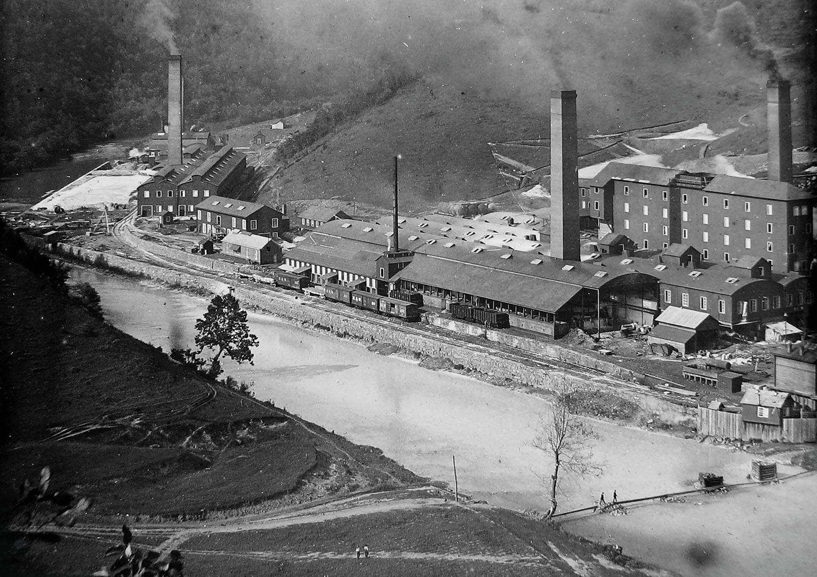 Une vieille photo en noir et blanc d'une grande usine chimique au bord d'une rivière, avec des cheminées qui crachent de la fumée noire dans l'air.