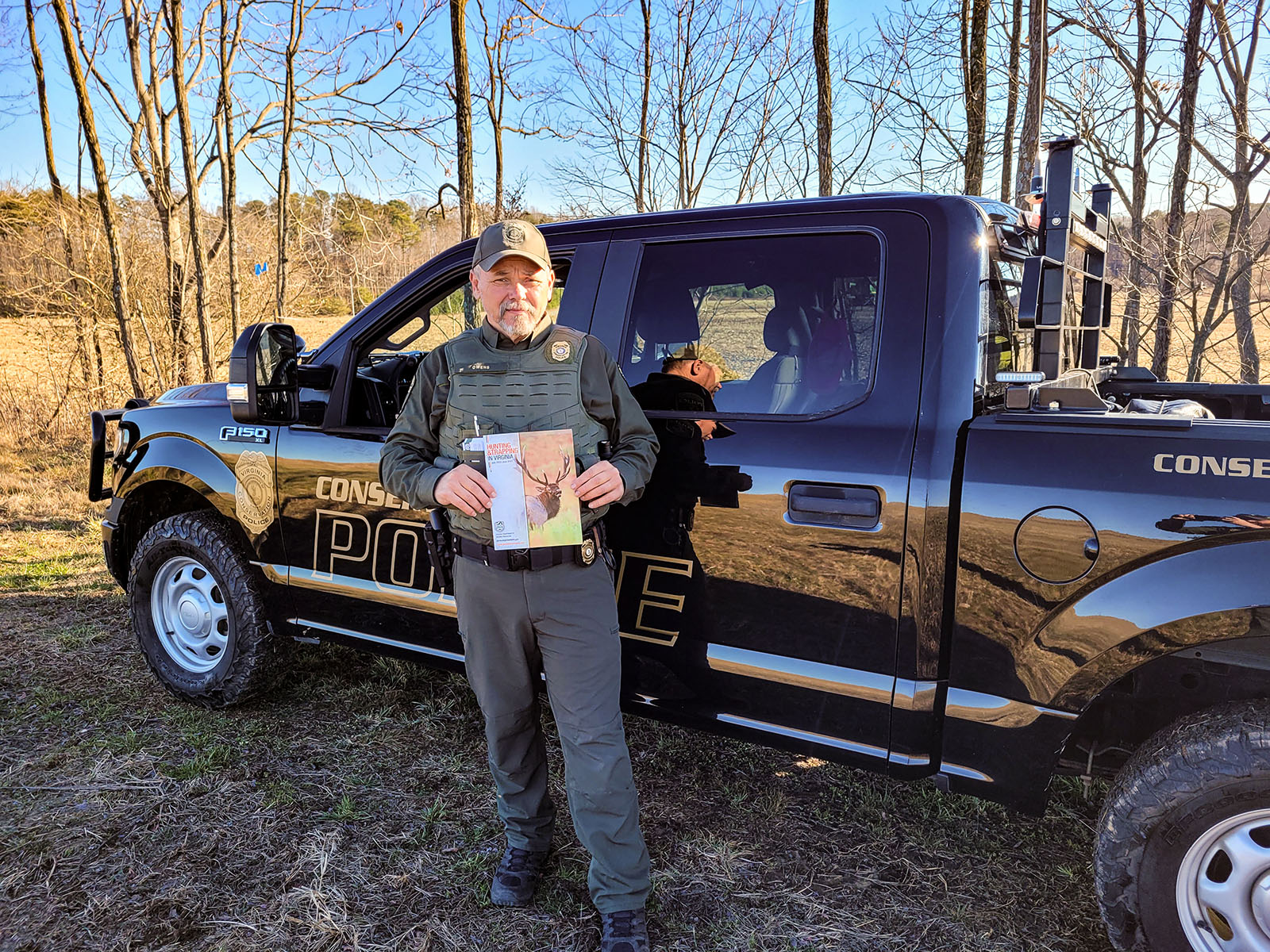 Une photo d'un agent de la police de la conservation en uniforme devant son camion, tenant une copie imprimée du livre 2023-24 Virginia Hunting and Trapping Regulations (Règlements de chasse et de piégeage de Virginie).