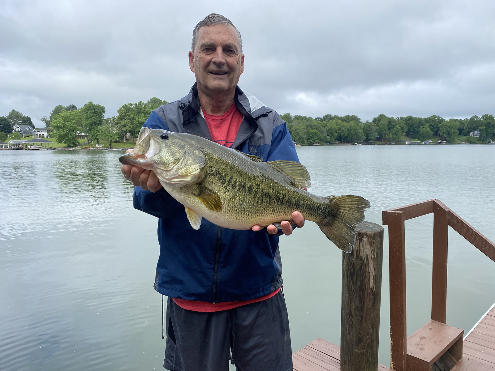 Photo d'un homme debout sur un quai devant un lac, tenant un très grand achigan à grande bouche.