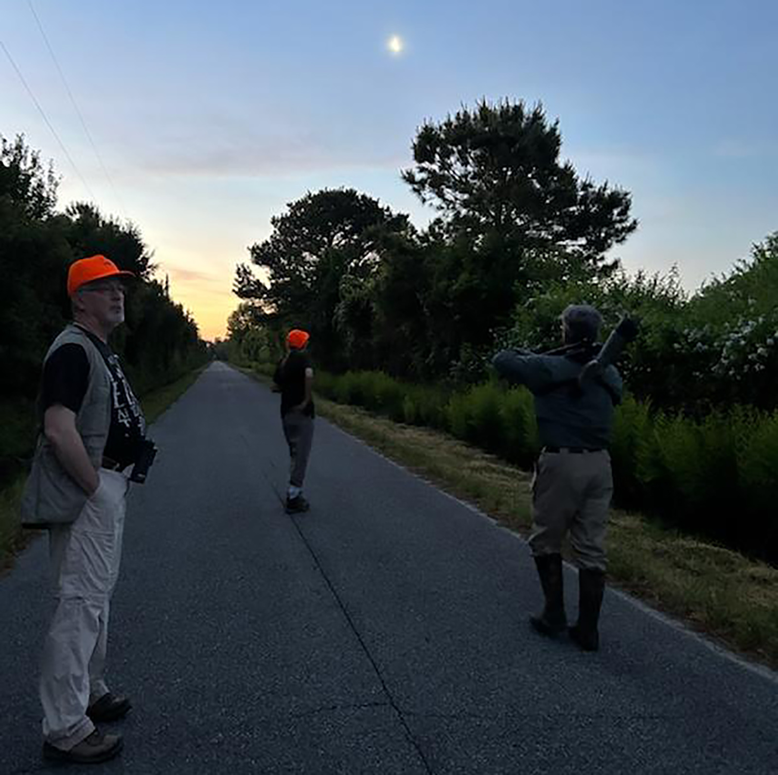 Une photo de trois personnes marchant le long d'une route dans une région isolée dans la lumière de l'aube avec la lune dans le ciel.