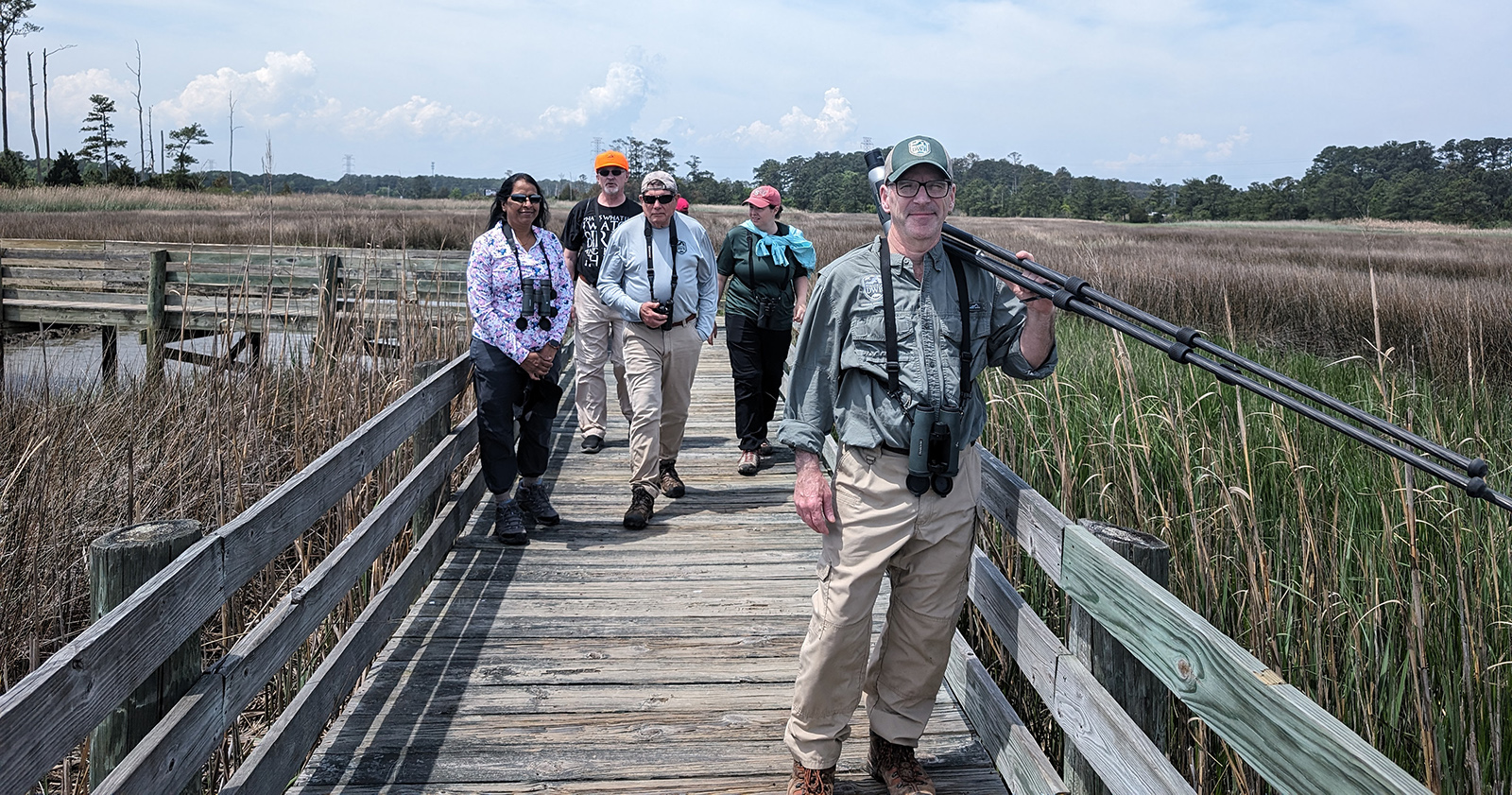 Une photo de cinq personnes avec des jumelles marchant sur une promenade en bois à travers une zone humide.