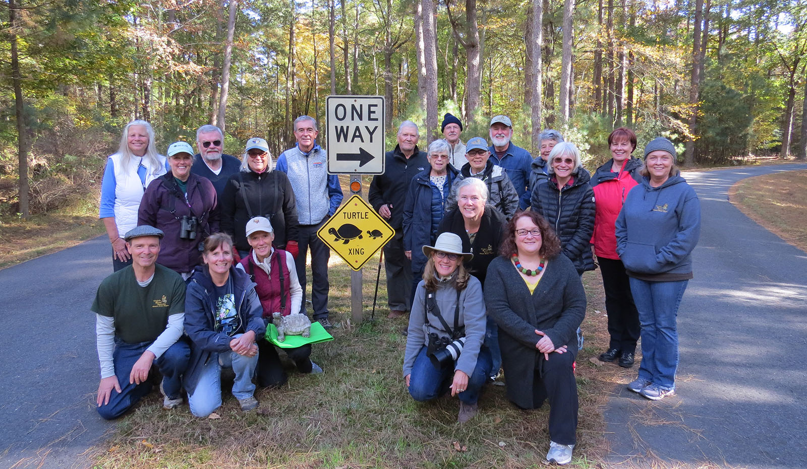 Un groupe de 20 personnes s'est rassemblé autour d'un panneau de passage pour tortues dans le terre-plein d'une petite route pavée, avec une personne agenouillée à l'avant avec une statue de tortue.