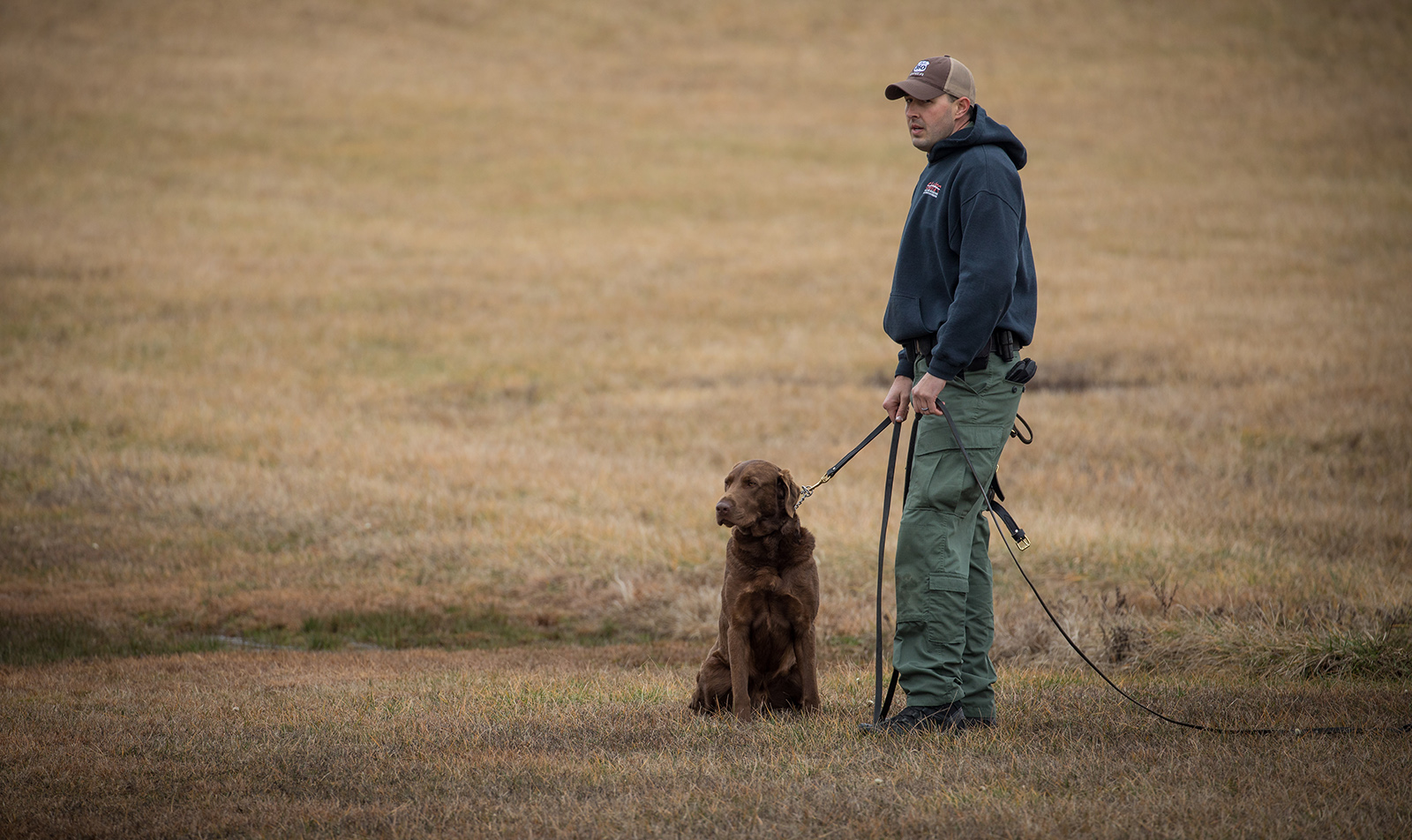 Une photo d'un homme debout, tenant la laisse d'un grand chien brun qui est assis tranquillement.