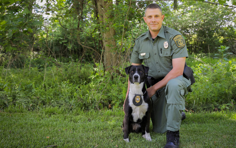 Une photo d'un homme en uniforme des forces de l'ordre agenouillé à côté d'un chien noir et blanc avec un badge autour du cou.