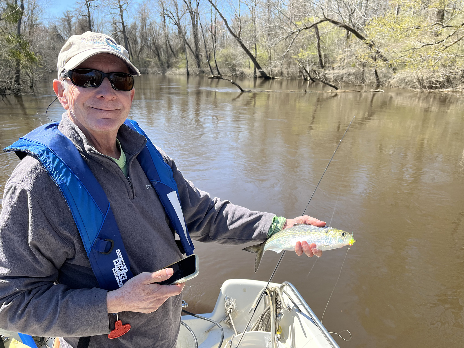 Un homme portant un gilet de sauvetage, debout sur un bateau dans une rivière et tenant un poisson alose brillant.