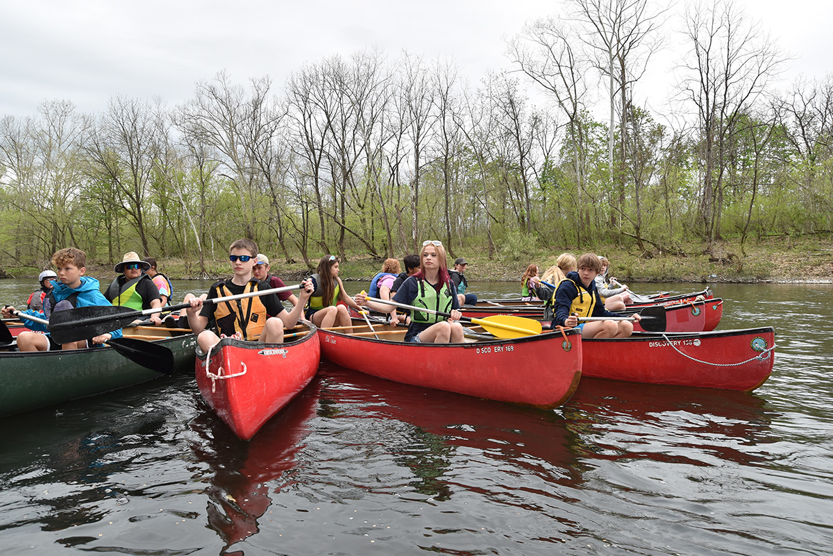 Une image de plusieurs étudiants dans des canoës rouges et verts et des gilets de sauvetage jaunes et orange sur l'eau de la rivière James.