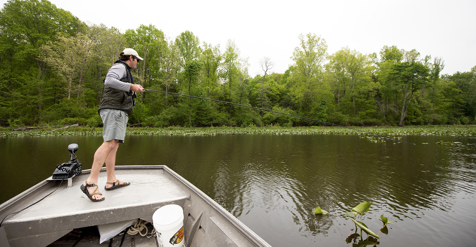 Un homme debout à l'avant d'un bateau à moteur, tenant une canne à pêche et regardant dans l'eau.