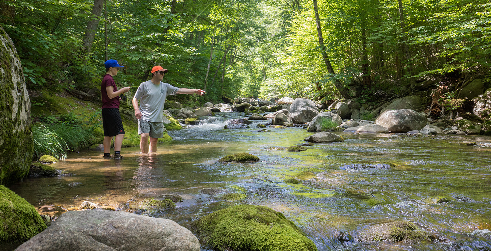 Une photo d'un homme pointant du doigt la rivière qui coule rapidement, tandis qu'un garçon se tient debout avec une canne à pêche et un moulinet.
