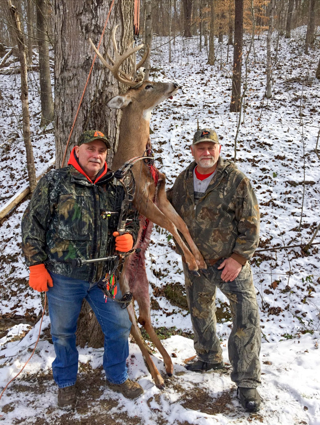 Une photo de deux hommes dans les bois avec de la neige au sol, debout près du corps d'un grand cerf de Virginie.