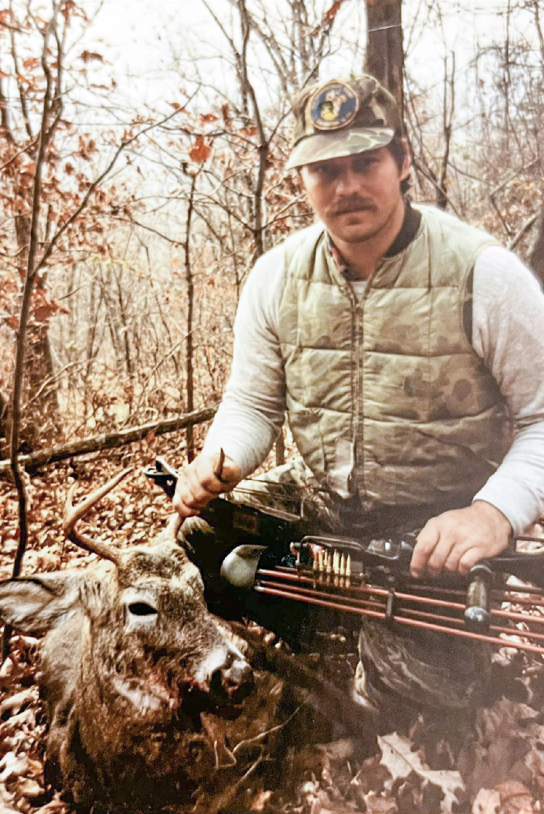 Une vieille photo d'un homme agenouillé dans les bois, tenant un arc à poulies et les bois d'un cerf de Virginie.
