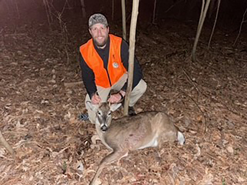Une photo d'un homme vêtu d'une veste orange vif, agenouillé derrière un petit cerf de Virginie au sol.