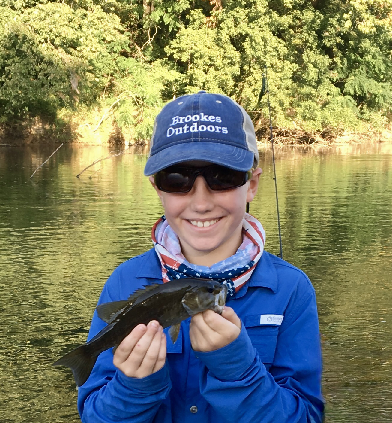 La photo d'un garçon tenant un achigan à petite bouche et souriant, avec une rivière derrière lui.