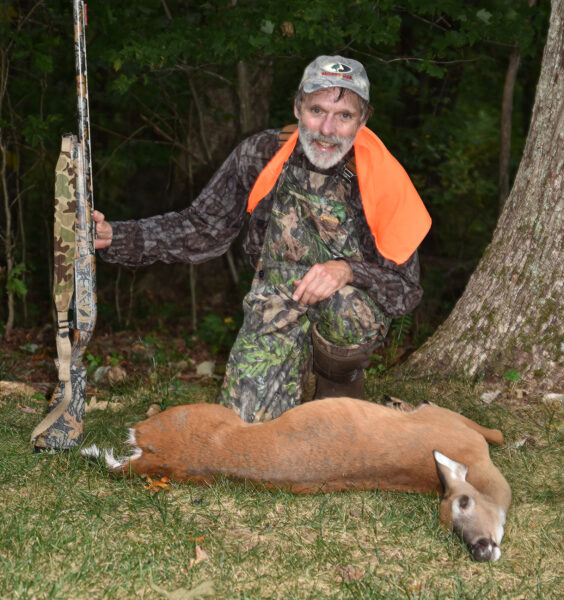Photo d'un homme en tenue de camouflage et orange tenant un fusil et agenouillé derrière une biche au sol.