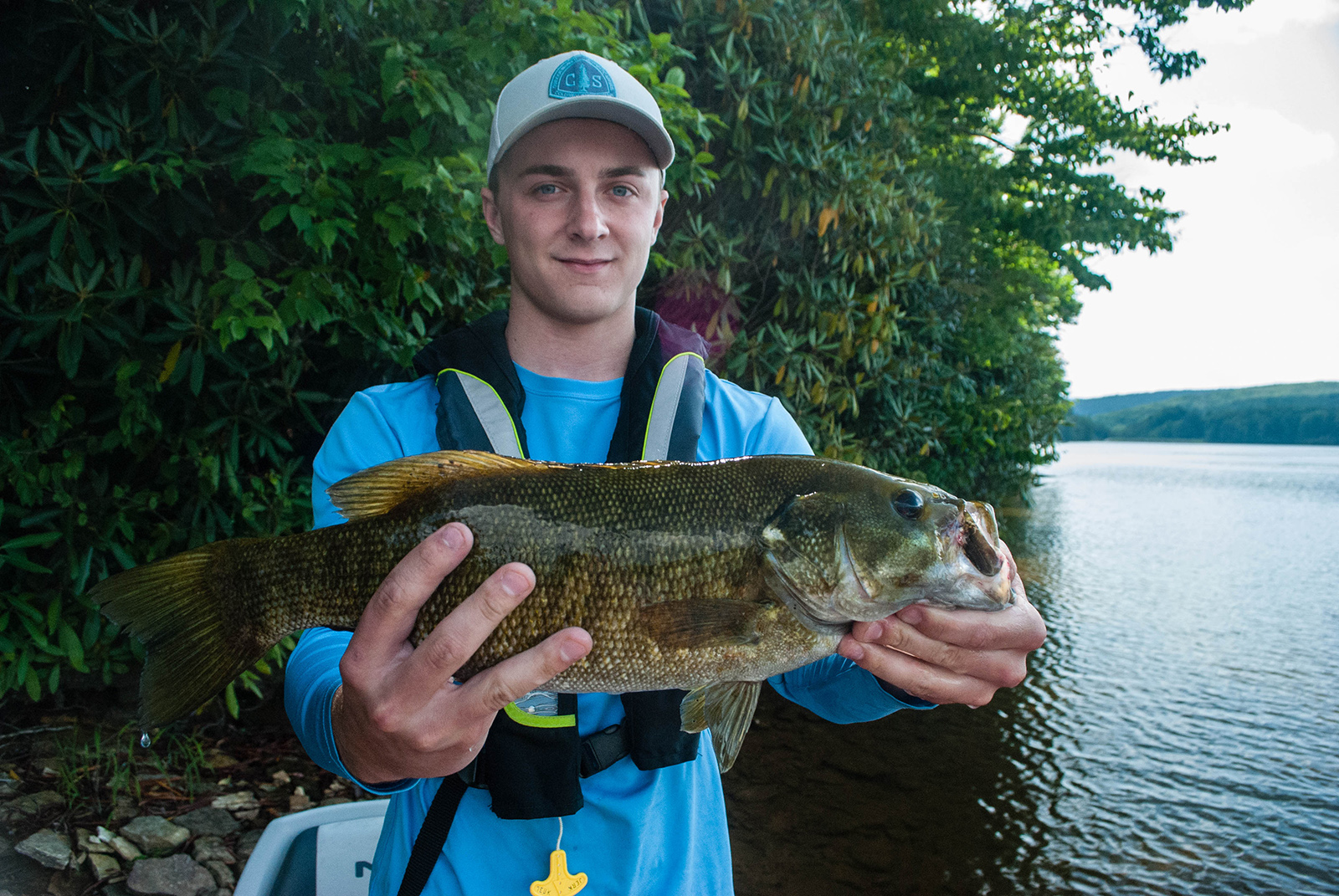 Photo d'un jeune homme portant un gilet de sauvetage et tenant un gros achigan à petite bouche, avec un lac en arrière-plan.