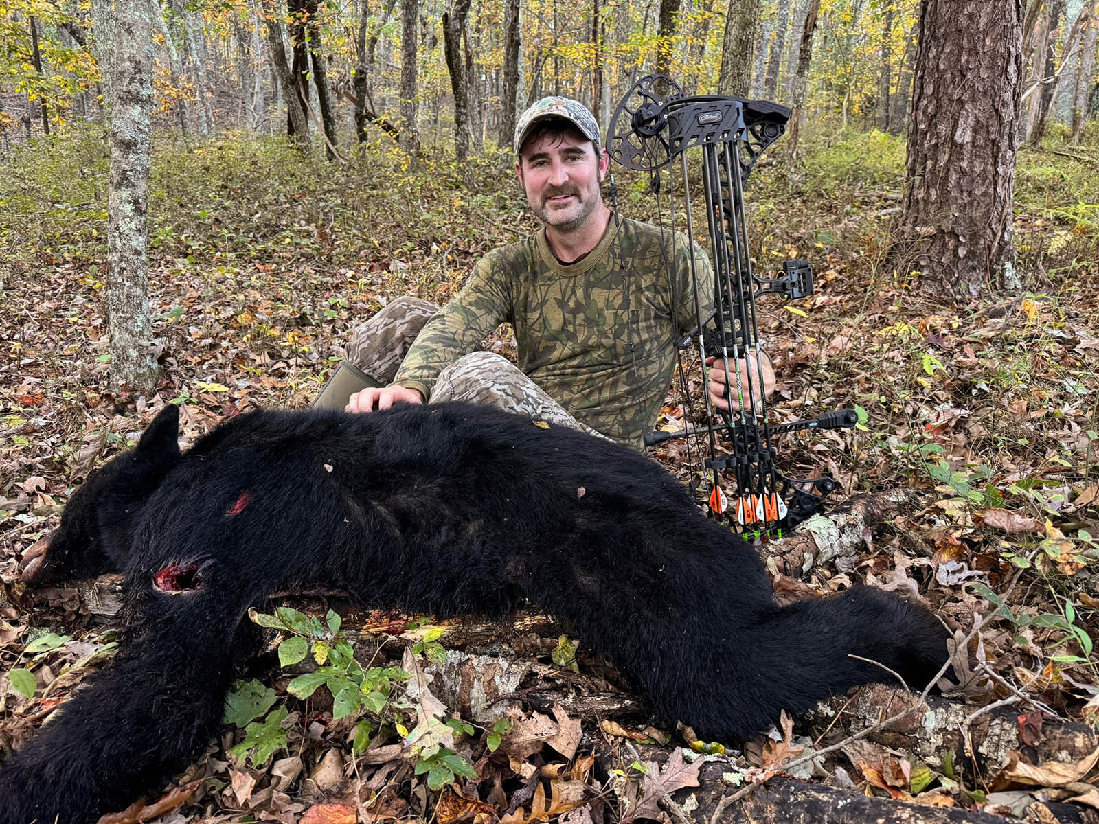 Une photo d'un homme portant un camouflage, assis dans les bois à côté d'un ours noir mort et tenant une arbalète.