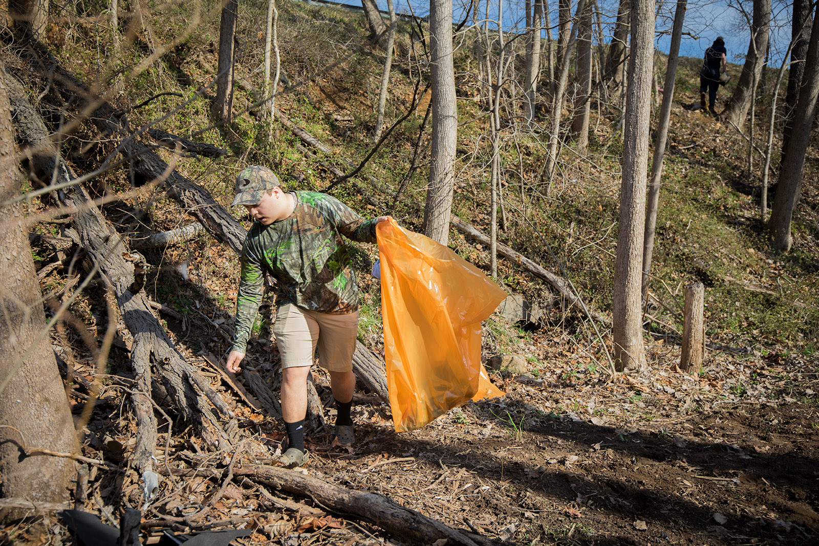 Image d'un homme en tenue de camouflage, muni d'un sac poubelle orange vif, ramassant des déchets dans une forêt. 