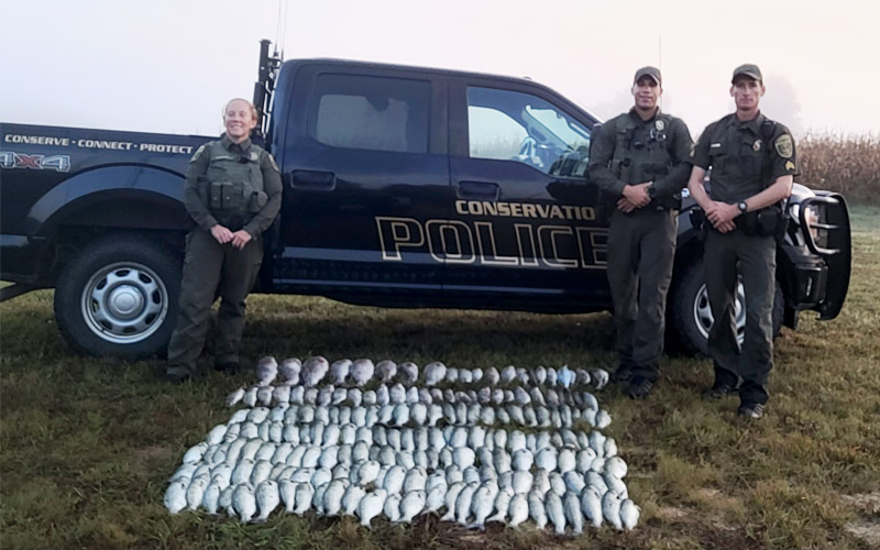Une photo de trois agents de la police de la conservation en uniforme se tenant devant un camion de la police de la conservation avec un grand nombre de poissons sur le sol devant eux.