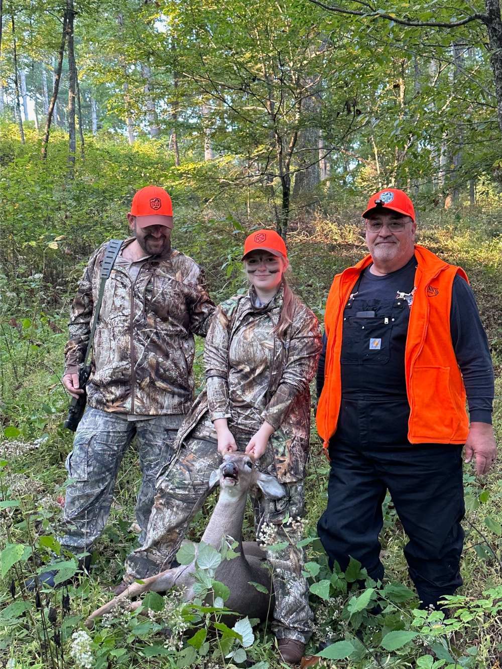 Photo d'une femme tenant la tête d'un cerf abattu, avec deux hommes à ses côtés. Ils sont dans les bois et tous habillés en camouflage et en orange flamboyant.