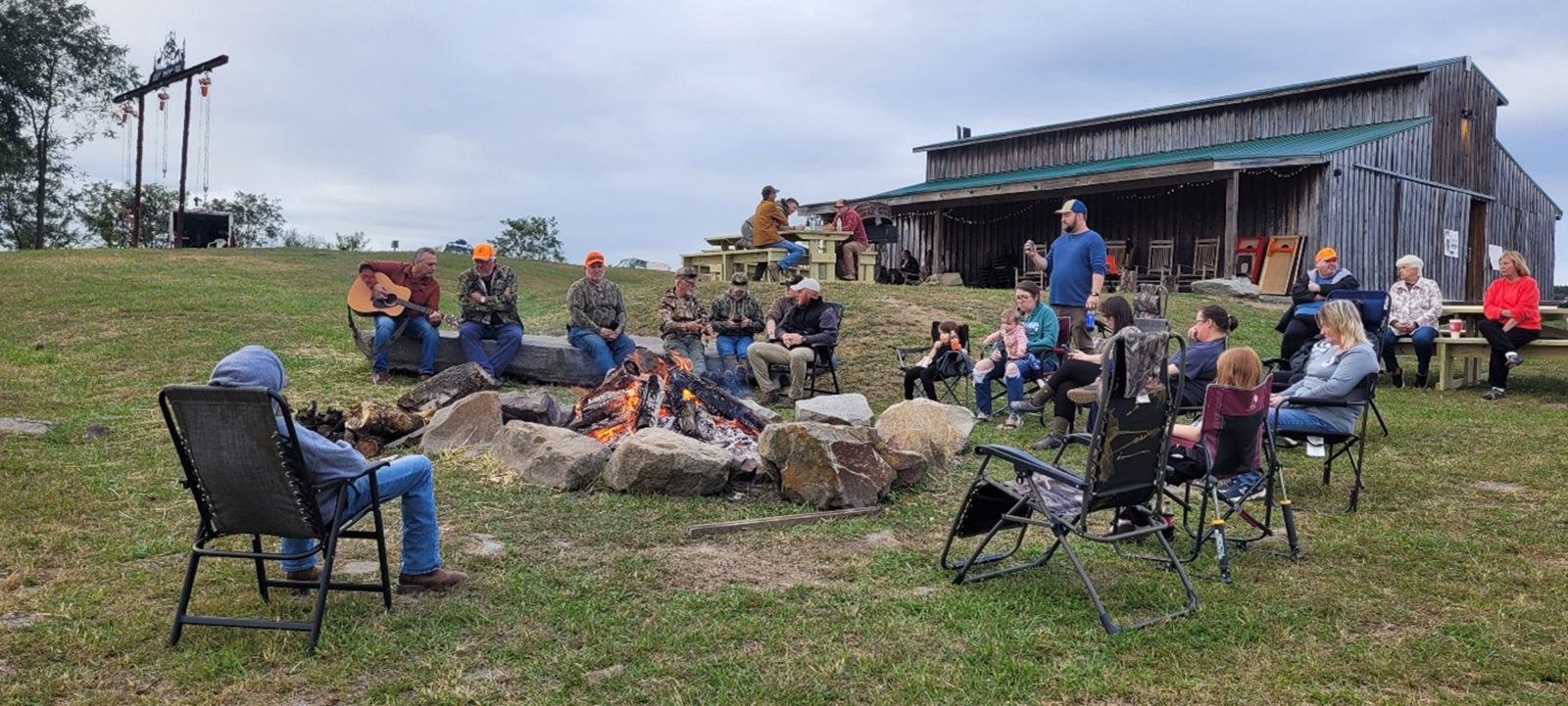 Photo d'un feu de camp avec de nombreuses personnes assises autour, avec une grange en arrière-plan.