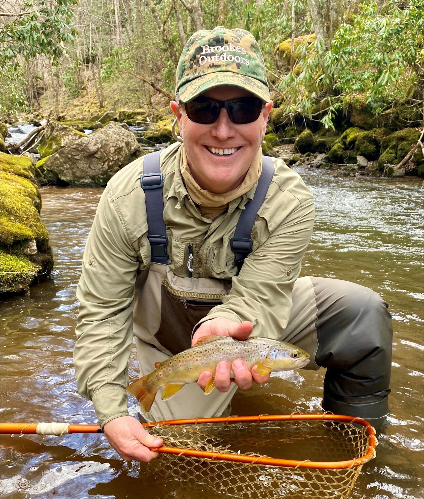 La photo d'un homme souriant, accroupi dans un beau ruisseau, tenant un filet de pêche et une truite brune colorée. 
