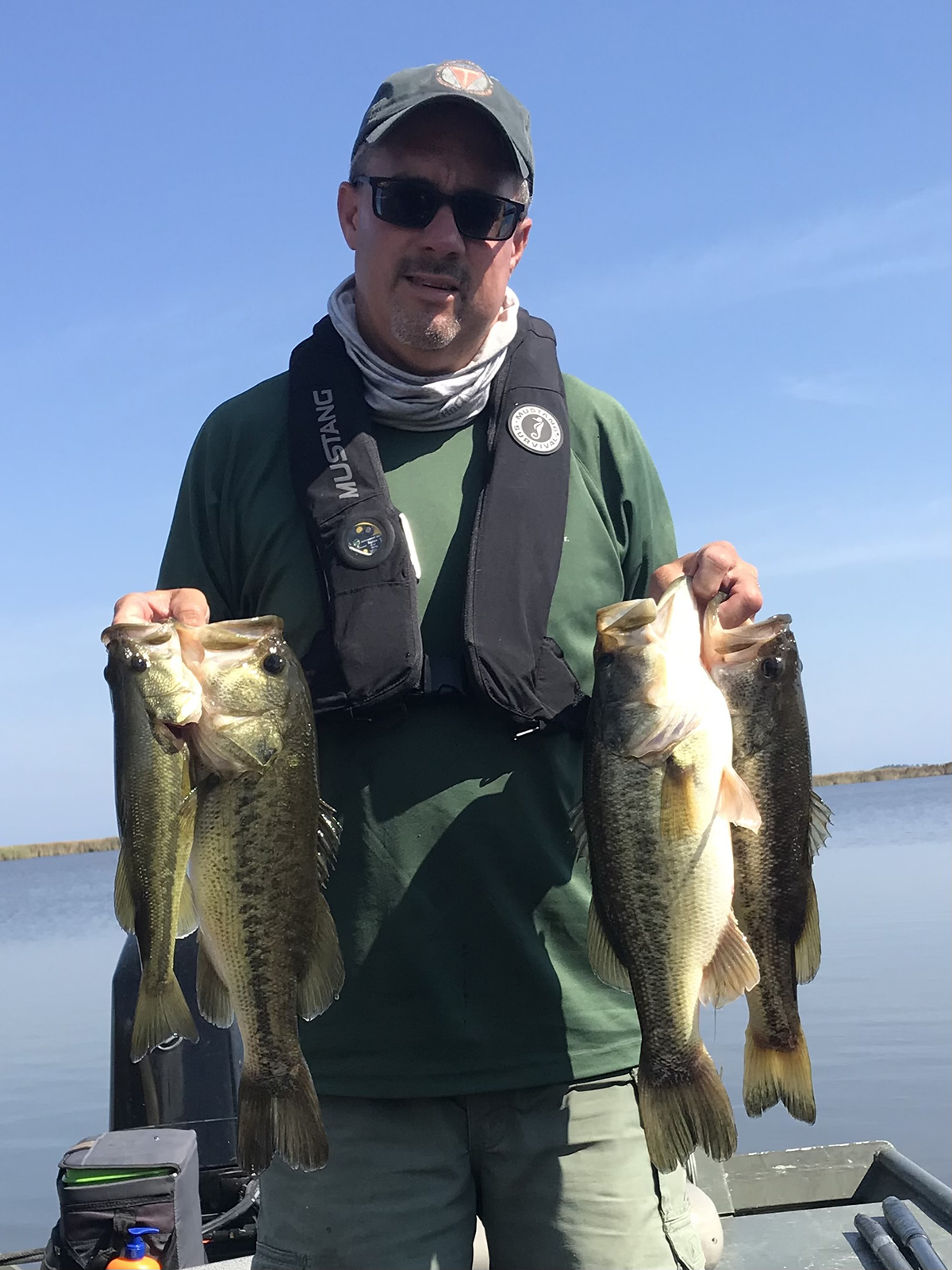 Un homme, Chad Boyce, tenant quatre achigans à grande bouche debout sur un bateau de pêche. Il porte un haut vert avec un gilet de sauvetage, des lunettes de soleil et une casquette de base-ball.