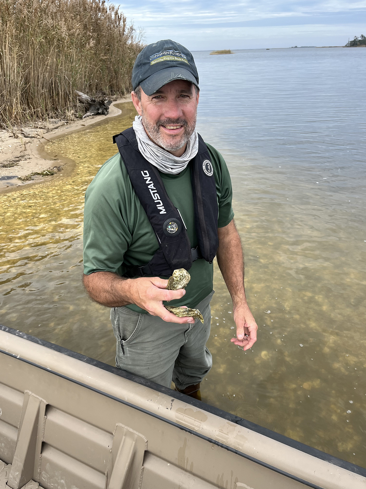 La photo d'un homme debout dans l'eau jusqu'aux genoux à côté d'un bateau, tenant des coquilles d'huîtres. 