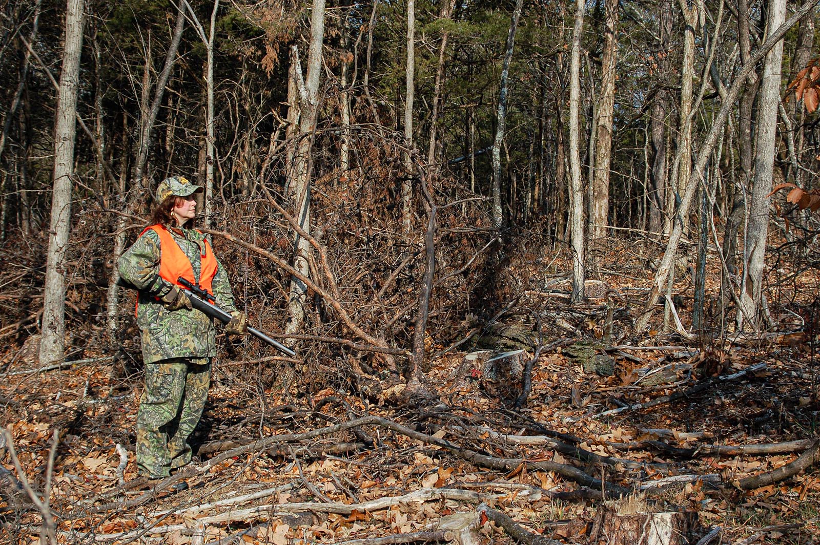 Photo d'un chasseur tenant un fusil, vêtu d'une tenue de camouflage et d'une tenue orange vif, au milieu d'arbres coupés dans une forêt.