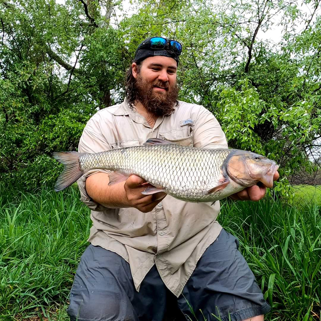 Photo d'un homme agenouillé sur la berge d'une rivière, tenant un grand poisson-chasseur.
