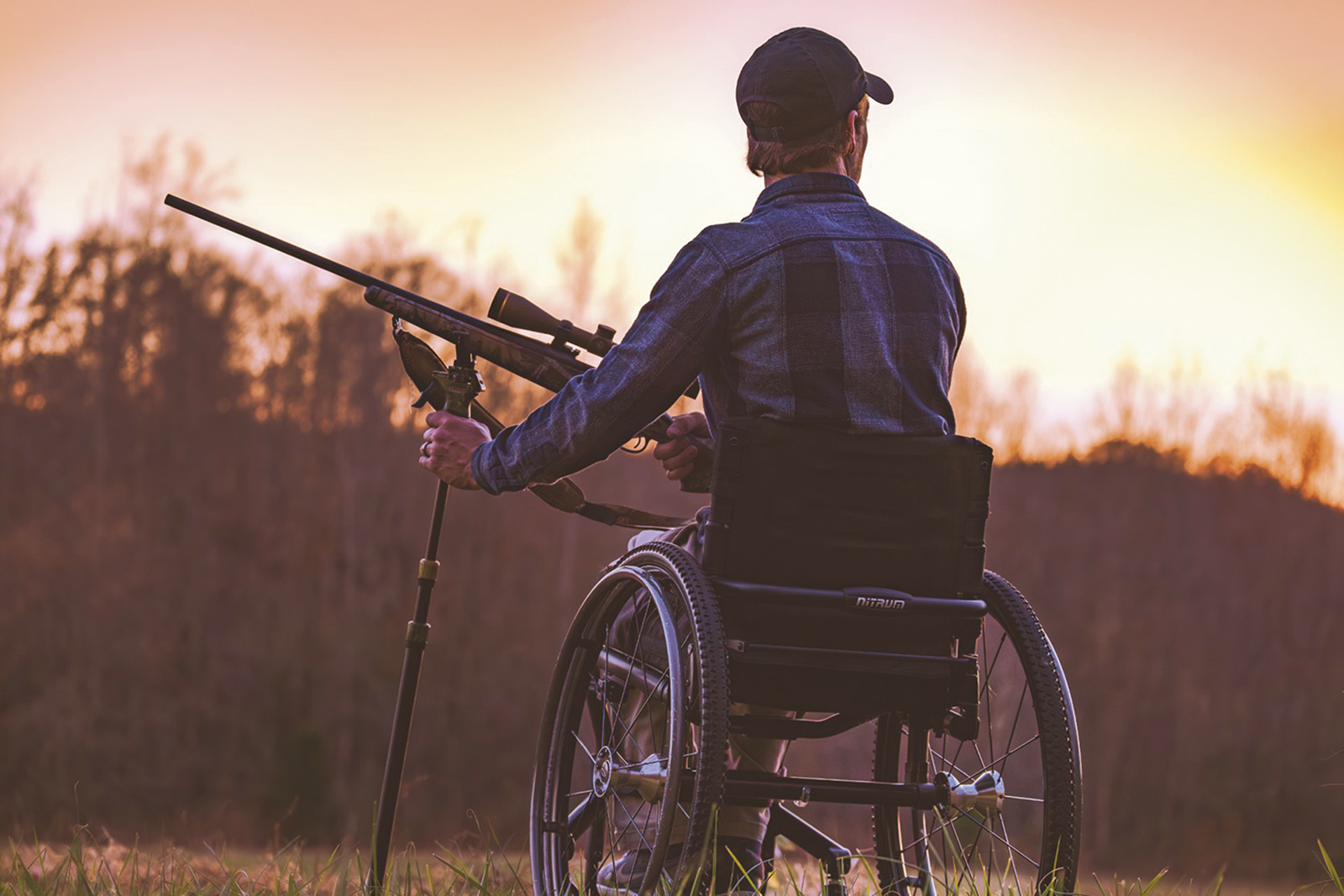 Une photo prise au crépuscule avec un chasseur en fauteuil roulant tenant un fusil et se détachant sur une ligne d'arbres.