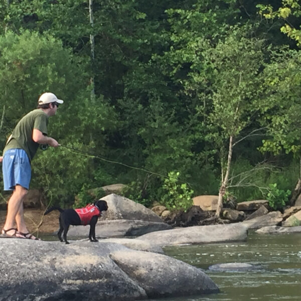 Image d'un homme pêchant avec un labrador noir portant un gilet de sauvetage rouge nommé Waffles sur un rocher dans la rivière James.